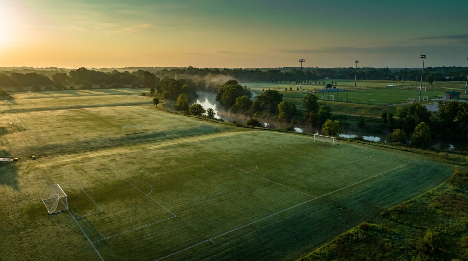 Aerial view of a soccer complex with six grass fields and one turf field at sunrise, goals lined up and empty